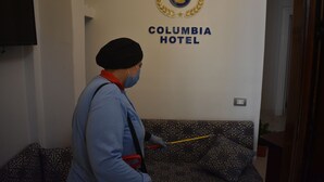 Ein Hotelmitarbeiter reinigt ein gemustertes Sofa in einem Zimmer mit einem an der Wand montierten Fernseher und einem Schild mit der Aufschrift 'COLUMBIA HOTEL'.