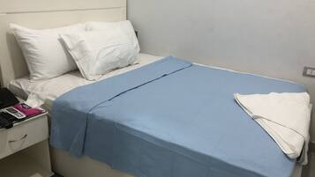 Una cama bien tendida con ropa de cama blanca y azul, una mesita de noche con un teléfono y un objeto pequeño encima, y un papel tapiz estampado en la pared.