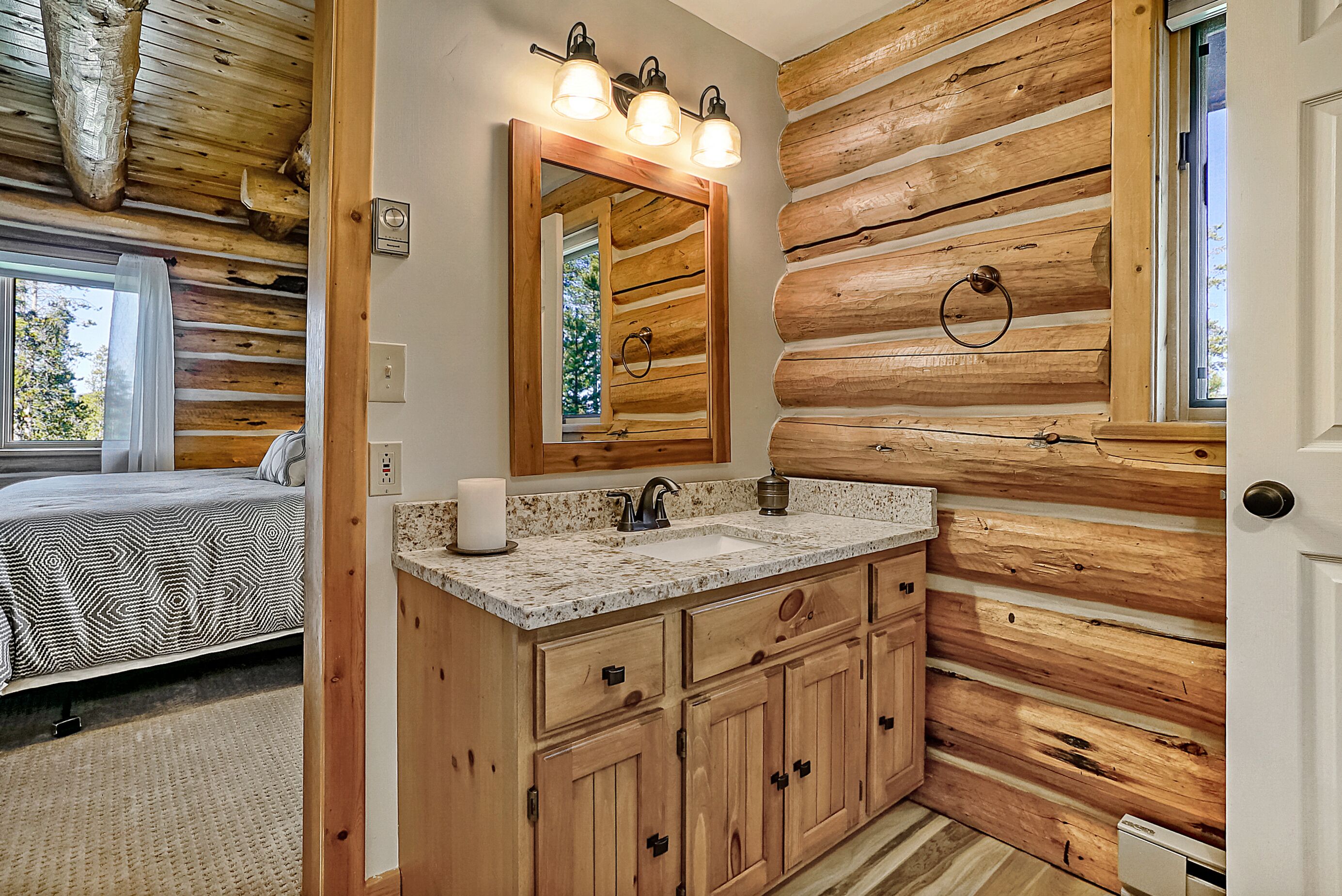 A log cabin bedroom with a bed, a bathroom with wooden cabinets, a sink, and a mirror with lights.