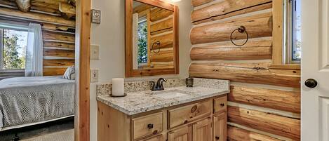 A log cabin bedroom with a bed, a bathroom with wooden cabinets, a sink, and a mirror with lights.