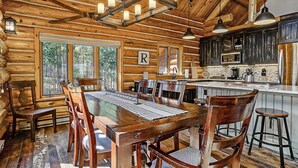 A rustic cabin interior with a wooden dining table, chairs, a kitchen area with modern appliances, and large windows offering a view of the forest.