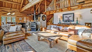 A spacious log cabin interior with a living room featuring leather sofas, a wooden coffee table, and a fireplace.