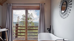 A bathroom with a bathtub, a wooden floor, a large window with curtains, and a view of trees and hills.