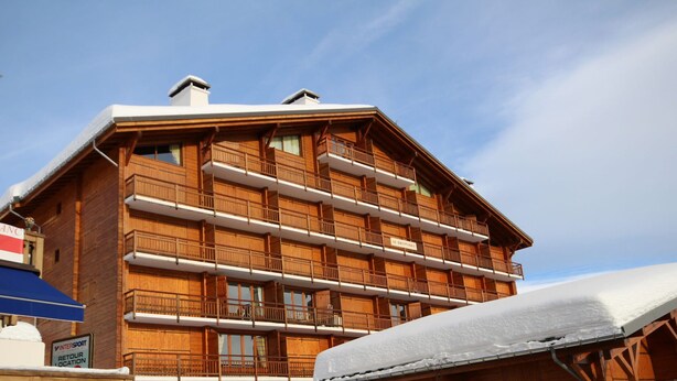 Ein mehrstöckiges Holzgebäude mit Balkonen, wahrscheinlich ein Hotel oder eine Lodge, mit einem Schild für "INTERSPORT" und einem Schild für "RETOUR LOCATION".