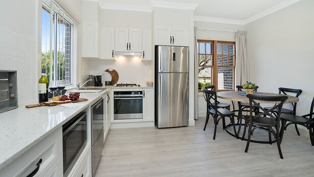 A modern kitchen with white cabinets, stainless steel appliances, a dining area with a wooden table and black chairs, and a large window with a view of greenery. - Accommodation Hunter - Adams St Maitland (Maitland)