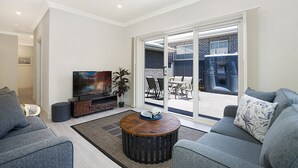 A modern living room with a grey sofa, a coffee table, a TV, and a sliding glass door leading to an outdoor patio. - Accommodation Hunter - Adams St Maitland (Maitland)