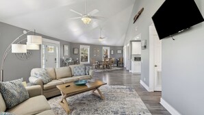 A spacious living room with a high ceiling, a ceiling fan, a flat-screen TV mounted on the wall, and a dining area in the background. - The Sand Dollar is a 3BD/2BA modern home w/pool, fenced in back yard, boat parking! (Mexico Beach)