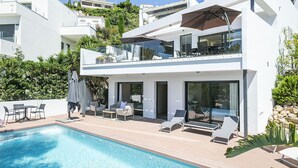 A modern two-story house with a swimming pool, outdoor seating, and a balcony with a large umbrella. - Noelia: Exclusive villa overlooking the sea and private pool. (L'Escala)
