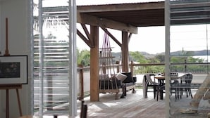 A spacious room with a large sliding glass door leading to an outdoor area with a wooden deck and a hammock. There is a dining table with chairs and a small table with a chair. - Haven Oceana (Emerald Beach)