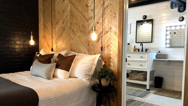 A bedroom with a herringbone-patterned wooden wall, a bed with white bedding and brown pillows, a small black side table with a plant, and a white dresser with a mirror above it. - 6 Bedroom/4.5 Bath/Across from Motown Museum (Detroit)