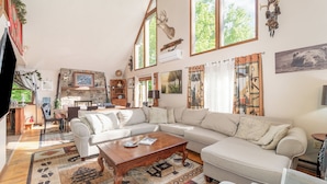 Amplia sala de estar con un sofá modular grande, una mesa de centro de madera y una chimenea. Varias ventanas proporcionan luz natural, y la habitación tiene una decoración rústica con vigas de madera y una chimenea de piedra.