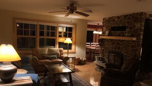 Un salon chaleureux avec une cheminée, un ventilateur de plafond et une grande fenêtre équipée de stores.