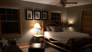 Une chambre confortable avec un lit en bois, une table de chevet avec une lampe, une fenêtre avec des stores et un ventilateur de plafond fixé au mur.