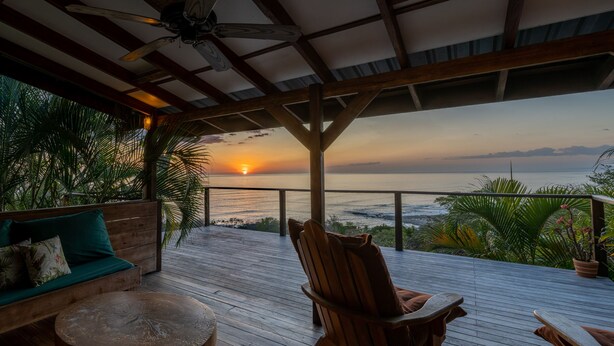 Una terraza de madera con vistas a la puesta de sol sobre el océano, un banco de madera y una mesa.