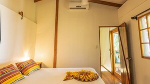 Un dormitorio con cama, almohadas, ventilador de techo, equipo de aire acondicionado, vigas de madera y una puerta que da a otra habitación.