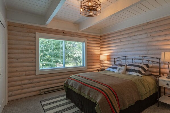 A log cabin bedroom with a large window, a bed with striped bedding, and a bedside lamp.