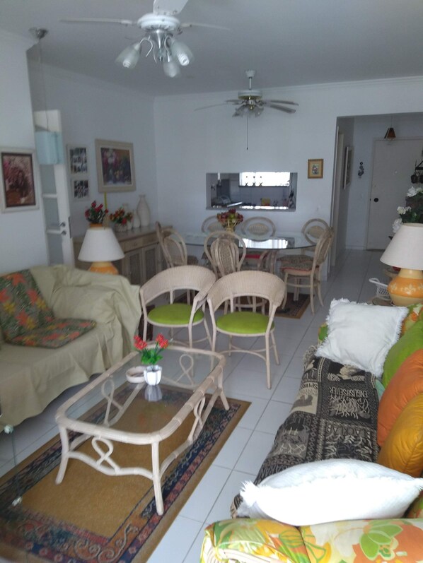Uma sala de estar com sofá, cadeiras, mesa de centro e mesa de jantar. Há um ventilador de teto, uma TV e uma porta ao fundo.