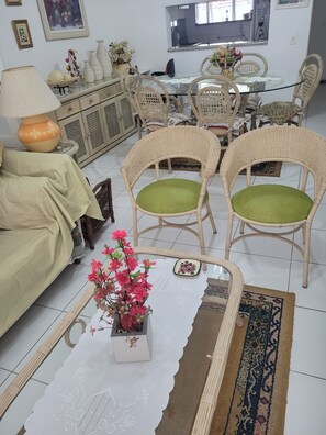 Uma sala de jantar com cadeiras de vime, uma mesa de vidro, um sofá com um travesseiro verde e um vaso de flores sobre uma mesa.