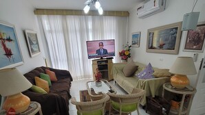 Una sala de estar con televisor, dos sofás, una mesa de centro, sillas de mimbre, un ventilador de techo y una ventana con cortinas.