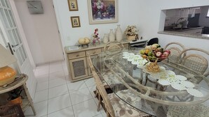 Un comedor con una mesa de vidrio rodeada de sillas de mimbre, piso de baldosas blancas y una cocina visible al fondo.
