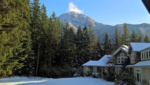 Una casa con tejado nevado, rodeada de frondosos árboles de hoja perenne y una montaña al fondo.