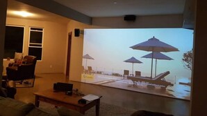 Una sala de estar con una pantalla grande que muestra una escena de playa, un sillón de cuero, una mesa de centro y una puerta que da al exterior.