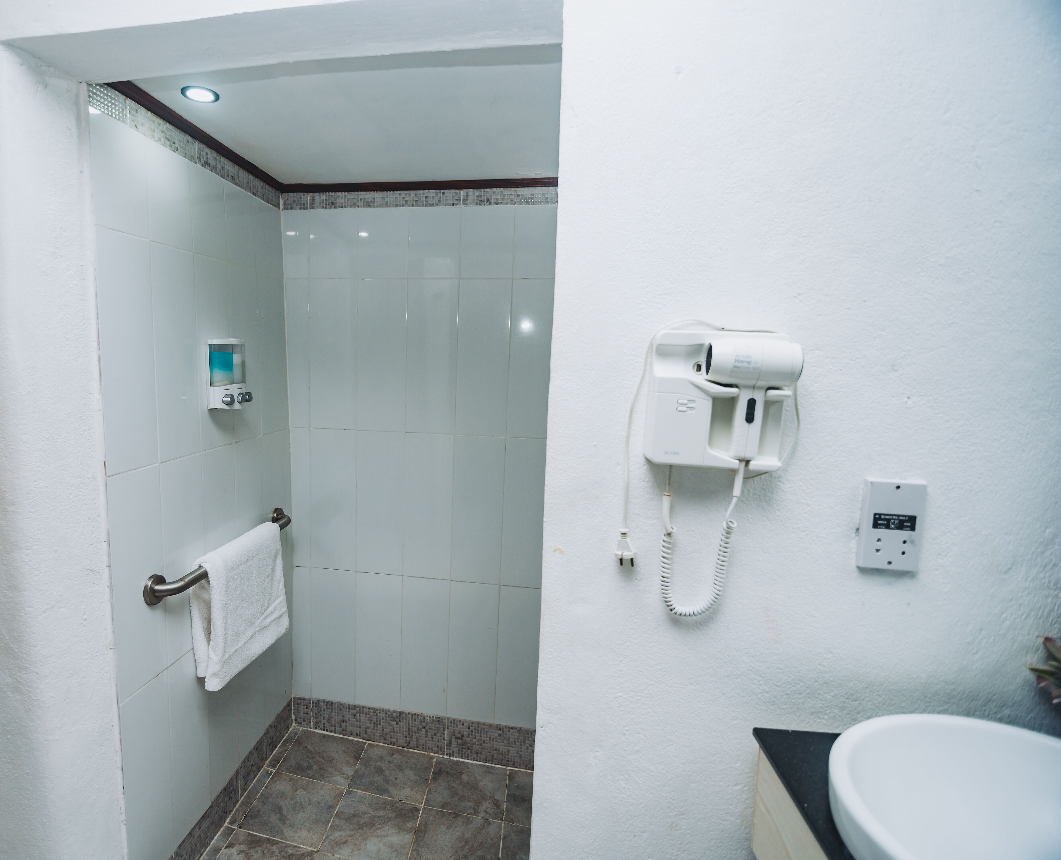 A bathroom with a white wall, a towel rack with a white towel, a hairdryer mounted on the wall, and a sink with a black countertop.