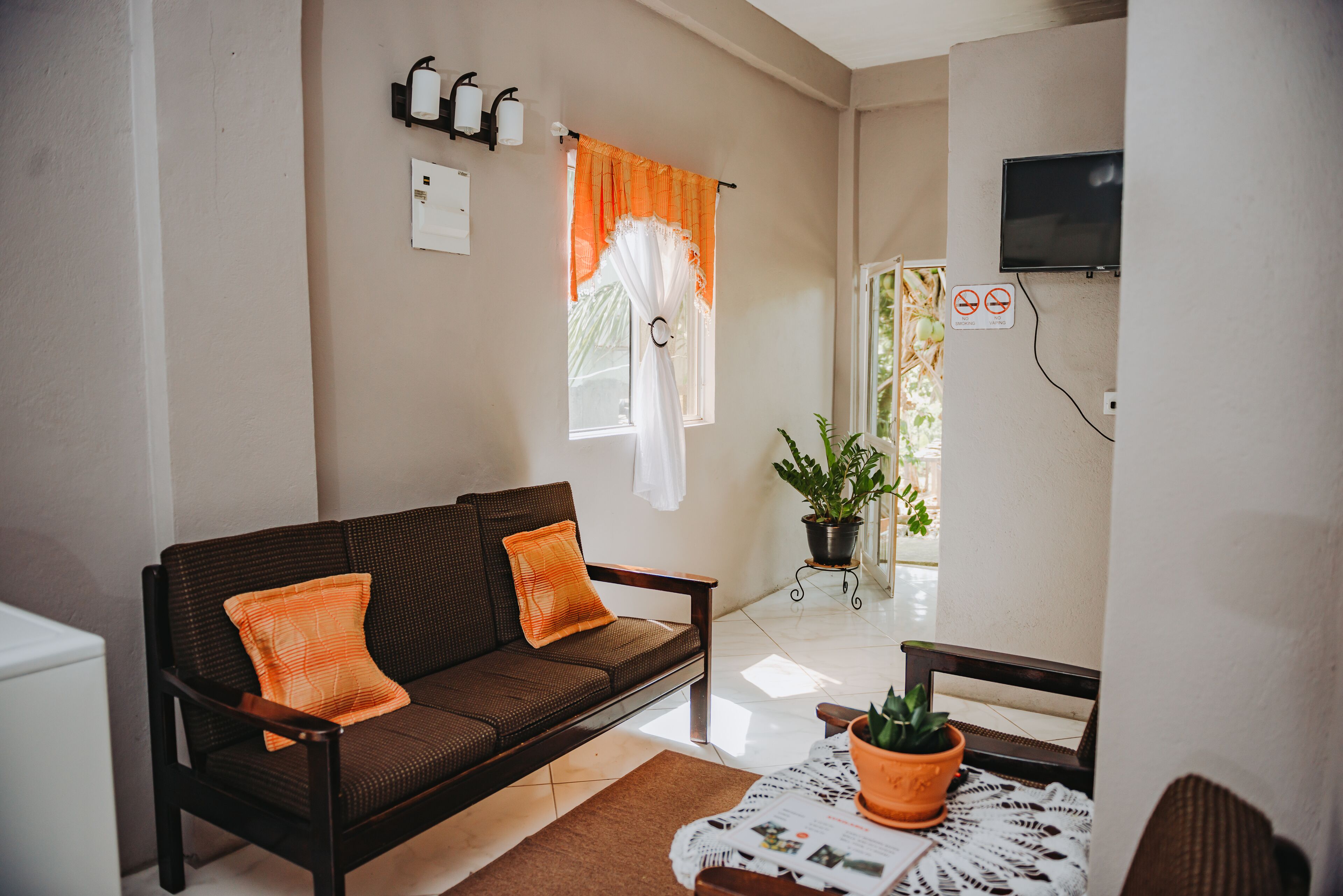Una sala de estar con un sofá marrón, cojines naranjas, una mesita con un periódico y una planta en maceta.