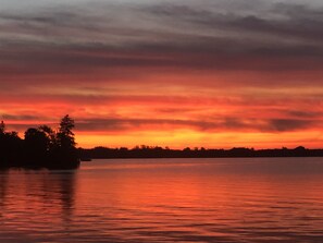 Un coucher de soleil sur un lac paisible, avec la silhouette d’arbres et une rive lointaine.