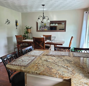 A dining area with a large granite table, wooden chairs, a chandelier, and a kitchen visible through a mirror.