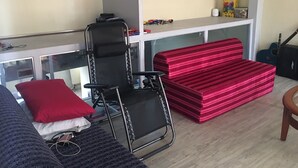 Ein Zimmer mit einem schwarzen Klappstuhl, einem rot gestreiften Sofa und einem hölzernen Stuhl. Es steht ein kleiner Tisch mit einem roten Kissen und einem weißen Kissen daneben. Im Hintergrund sind eine Gitarre und ein Lautsprecher zu sehen.