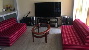 Ein Wohnzimmer mit Fernseher, Lautsprechern und einer Gitarre. Zwei gestreifte Sofas und ein Couchtisch aus Holz sind vorhanden.