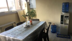 Ein Essbereich mit einem Tisch, der mit einer gemusterten Tischdecke bedeckt ist, Stühlen, einem Wasserspender und einer großen Wasserkaraffe.