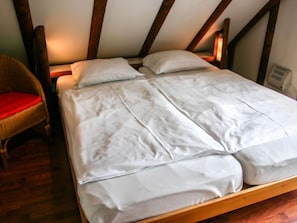 Une chambre à coucher avec un lit en bois, une literie blanche et un fauteuil en osier agrémenté d’un coussin rouge.