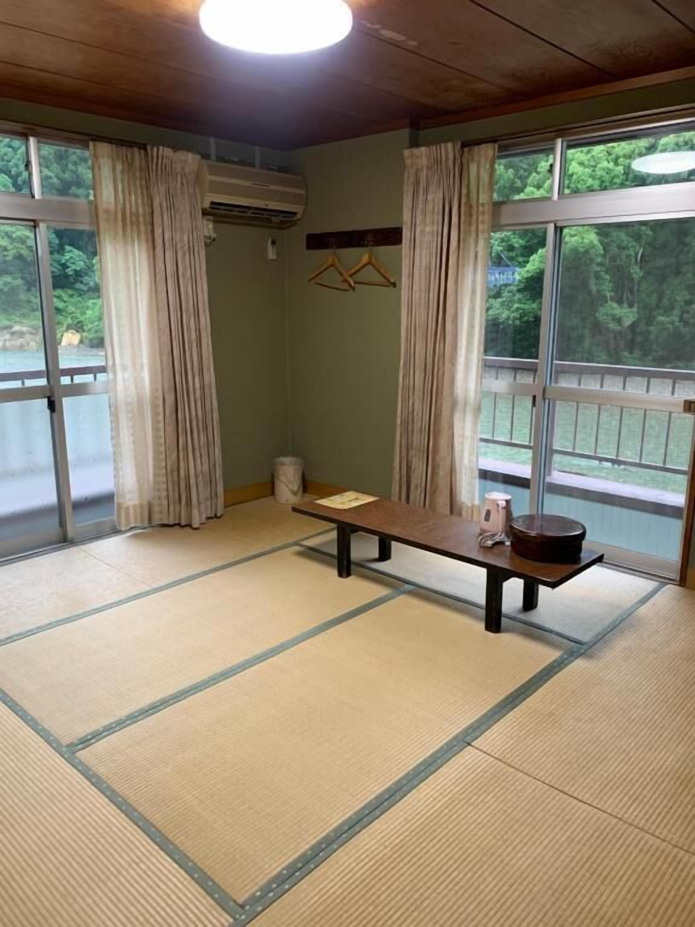Una habitación con suelo de tatami, una mesa de madera, aire acondicionado colgante y vistas a zonas verdes a través de ventanas amplias.
