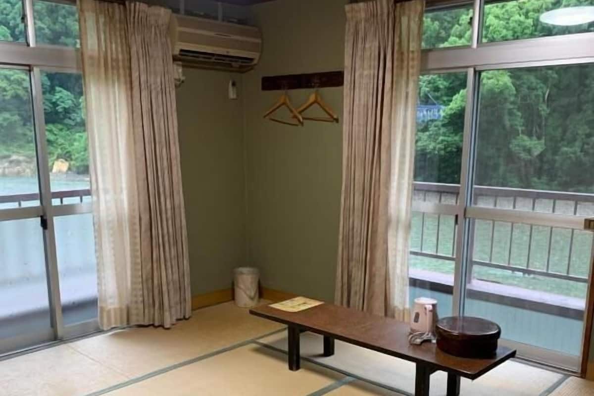 A room with tatami flooring, a wooden table, a hanging air conditioner, and a view of greenery through large windows.