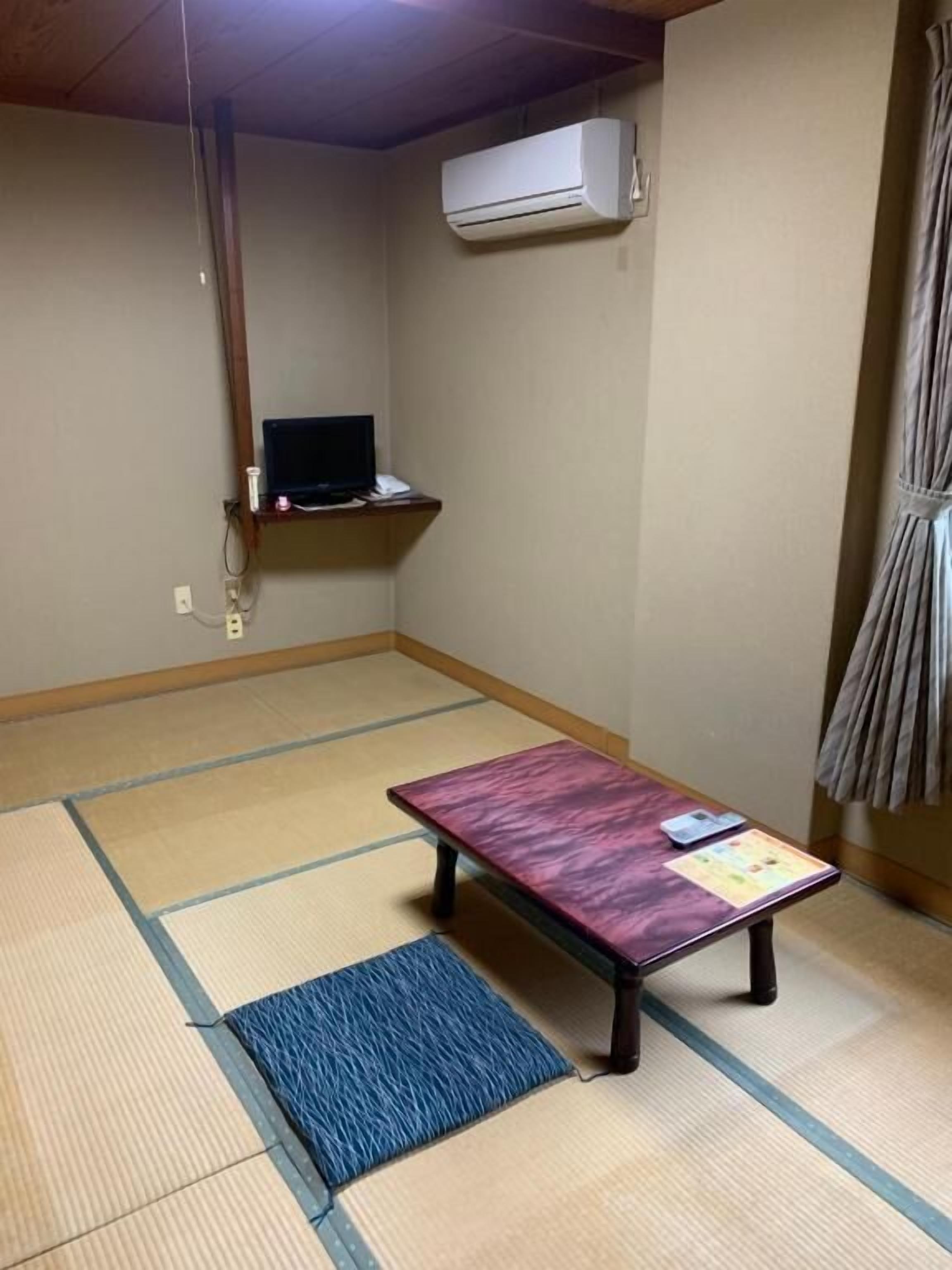 Ein Raum mit Tatami-Matten, einem Tatami-Tisch, einem kleinen Regal mit einem Fernseher und einer Klimaanlage an der Wand.