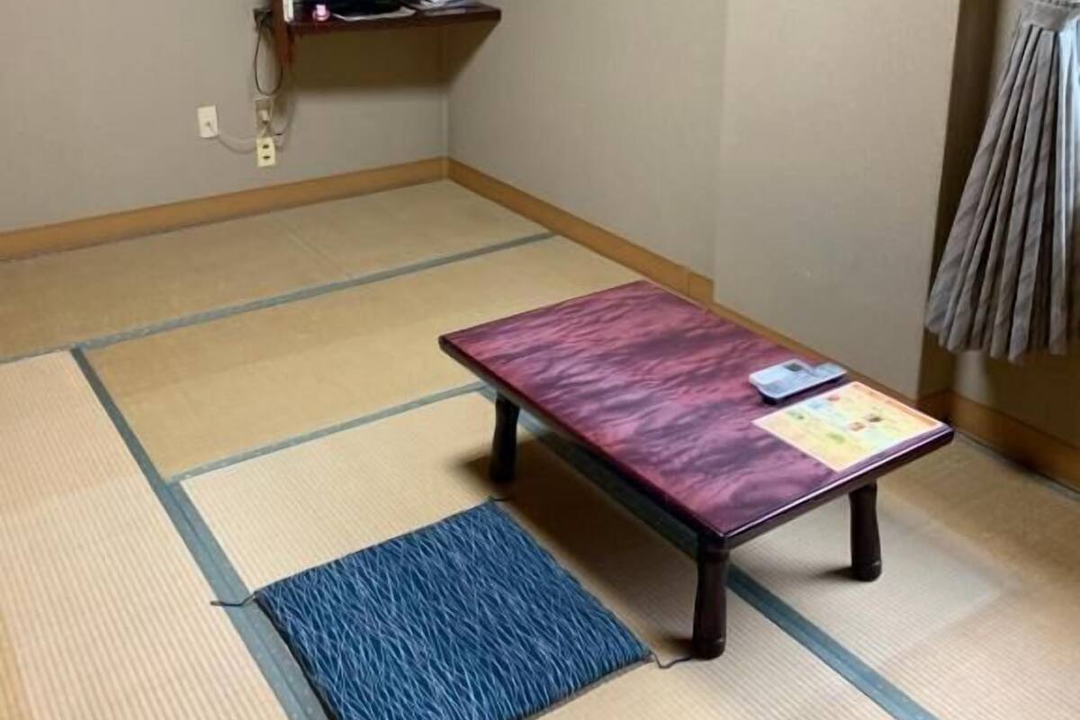 Une pièce avec un sol recouvert de tatamis, une table basse en tatami, une petite étagère avec un téléviseur et un appareil de climatisation fixé au mur.