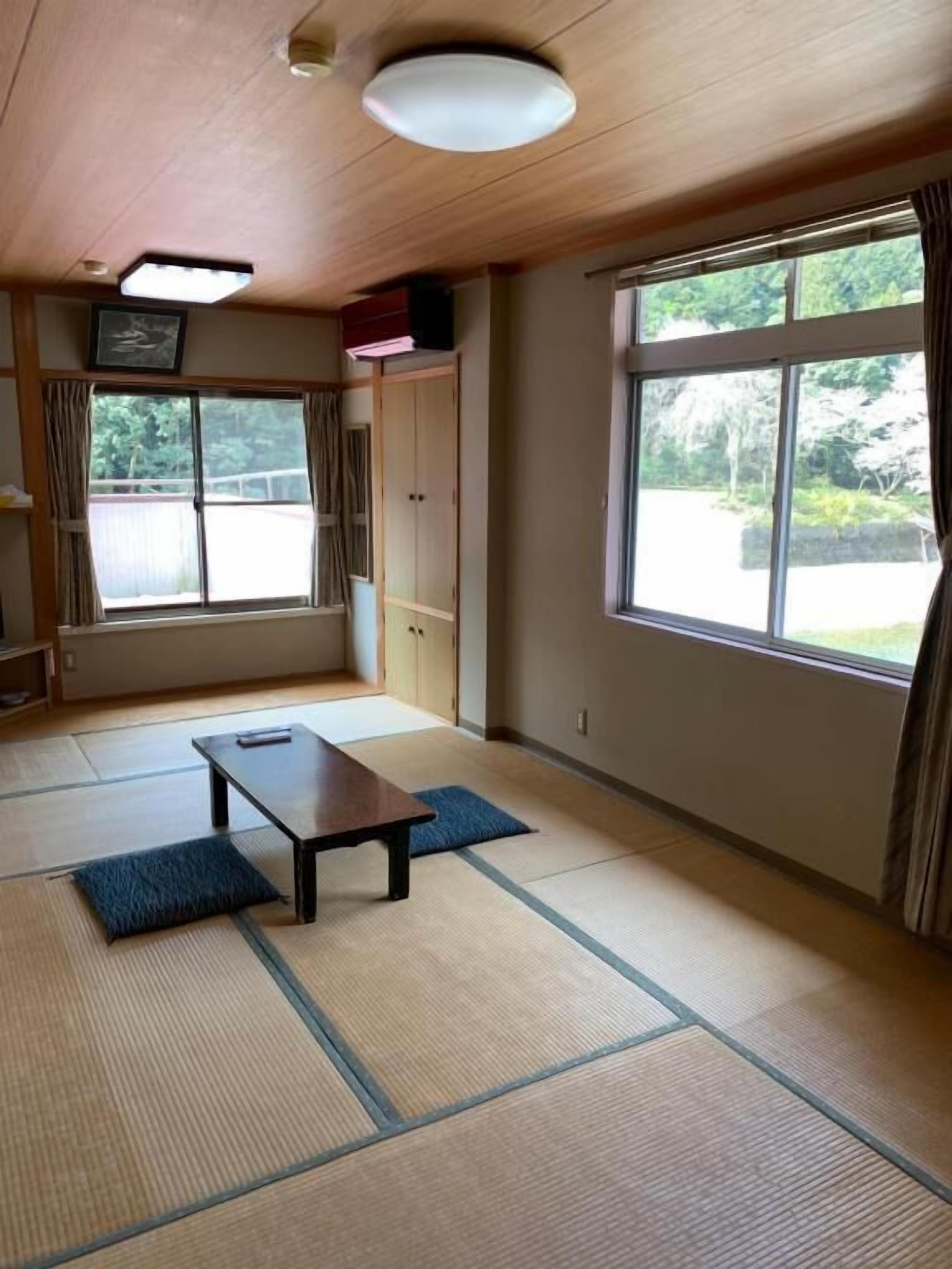 Una habitación con suelo de tatami, una mesa baja, un armario y ventanas grandes con cortinas.
