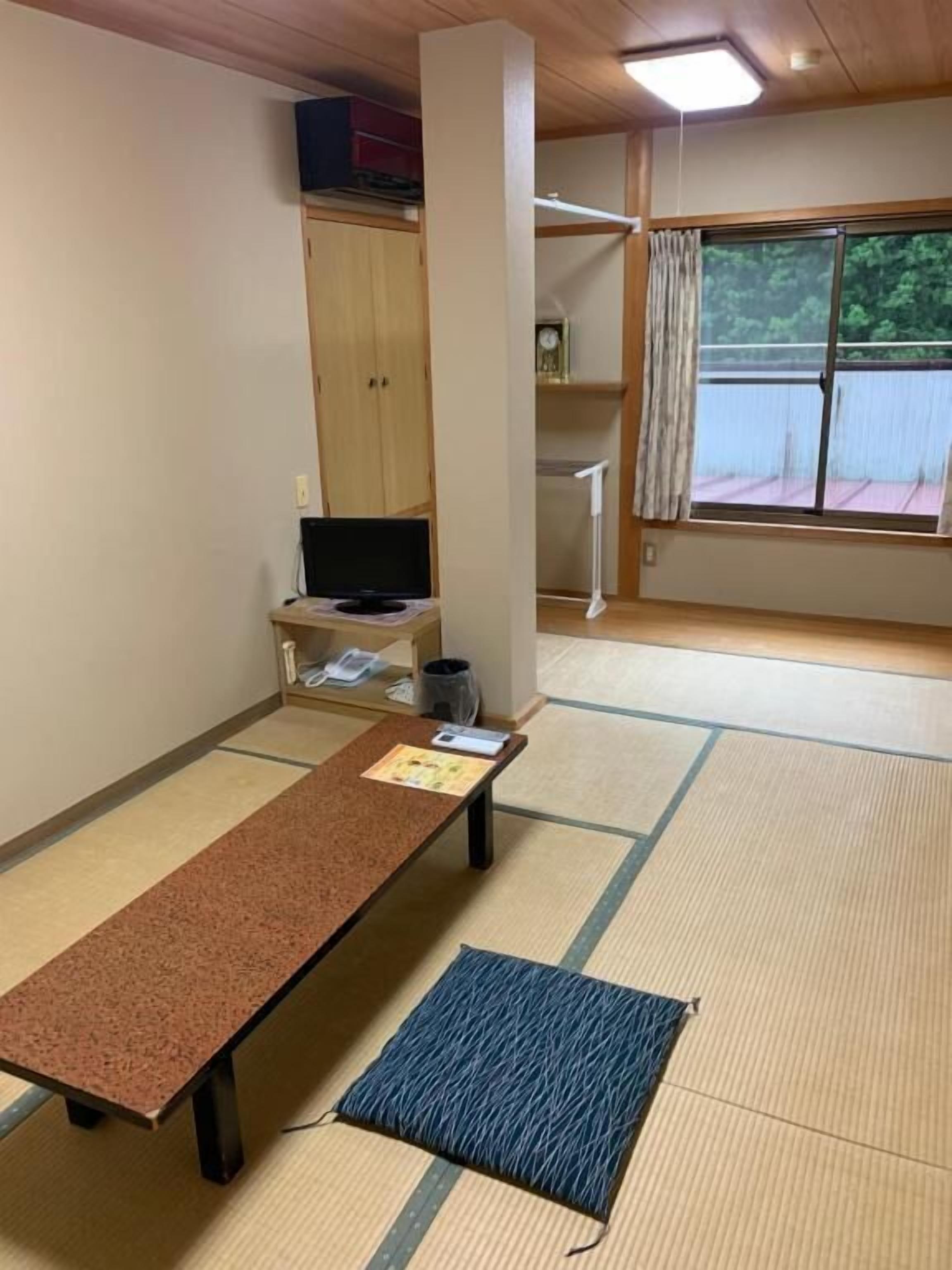 Una habitación con suelo de tatami, una mesa baja de tatami, un televisor sobre un mueble bajo, un armario y una ventana con vistas a zonas verdes.