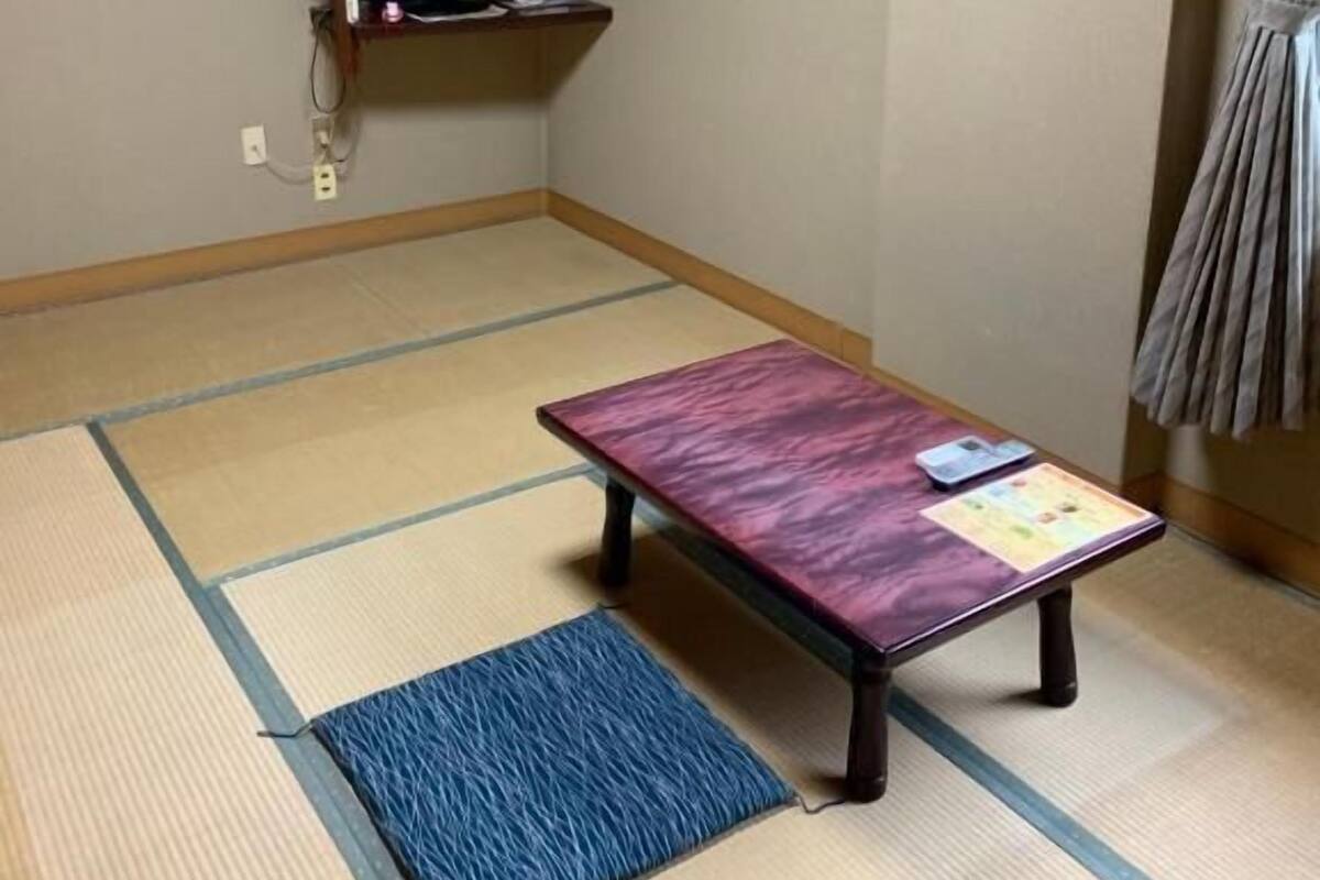 Une pièce avec un sol recouvert de tatamis, une table, une étagère avec un téléviseur et un climatiseur.