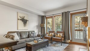 Una sala de estar moderna con sofá, sillones, una mesa de centro y una mesa de comedor. Hay un ventanal con cortinas, un cuadro de un alce en la pared y piso de madera.