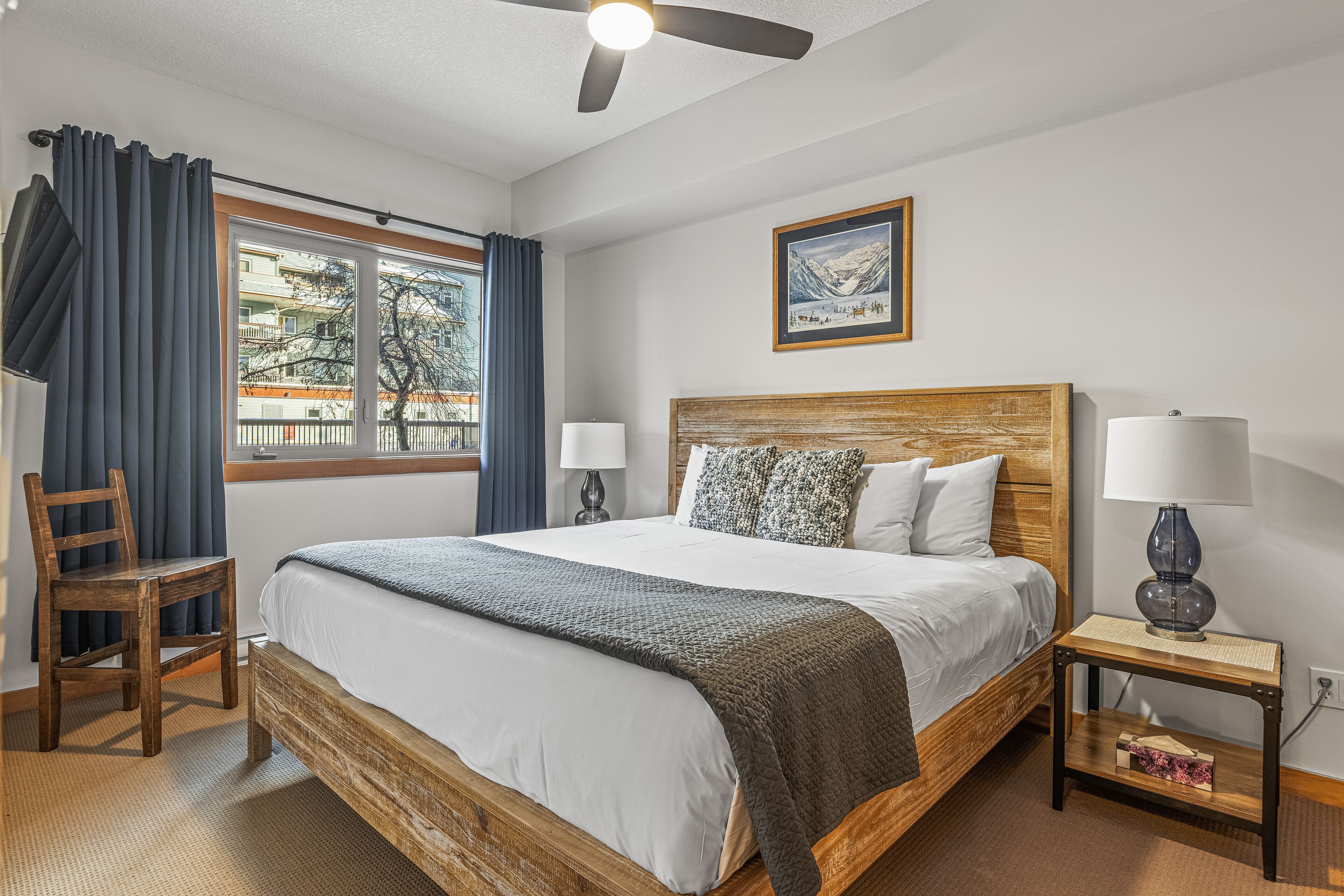 Un dormitorio con una cama grande, cabecera de madera, dos mesitas de noche con lámparas, una silla, una ventana con cortinas y un cuadro enmarcado en la pared.