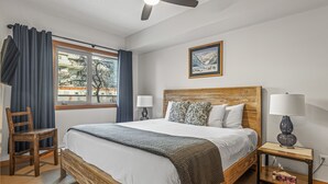 Un dormitorio con una cama grande, cabecera de madera, dos mesitas de noche con lámparas, una silla, una ventana con cortinas y un cuadro enmarcado en la pared.