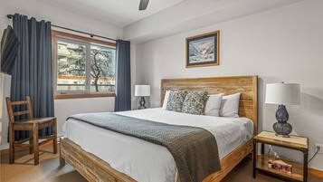 Un dormitorio con una cama grande, cabecera de madera, dos mesitas de noche con lámparas, una silla, una ventana con cortinas y un cuadro enmarcado en la pared.