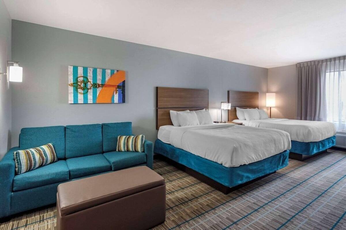 Habitación de hotel con sofá, dos camas, piso alfombrado y un cuadro en la pared.