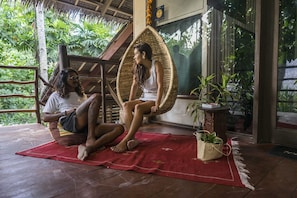 Two people sitting on a red mat in a room with a thatched roof, a wicker chair, and a potted plant.