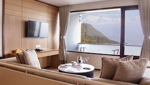 Ett modernt hotellrum med ett stort fönster som erbjuder utsikt över bergen, en platt-TV som är monterad på väggen, en mysig sittgrupp med ett bord för två och en väggpanel i trä.