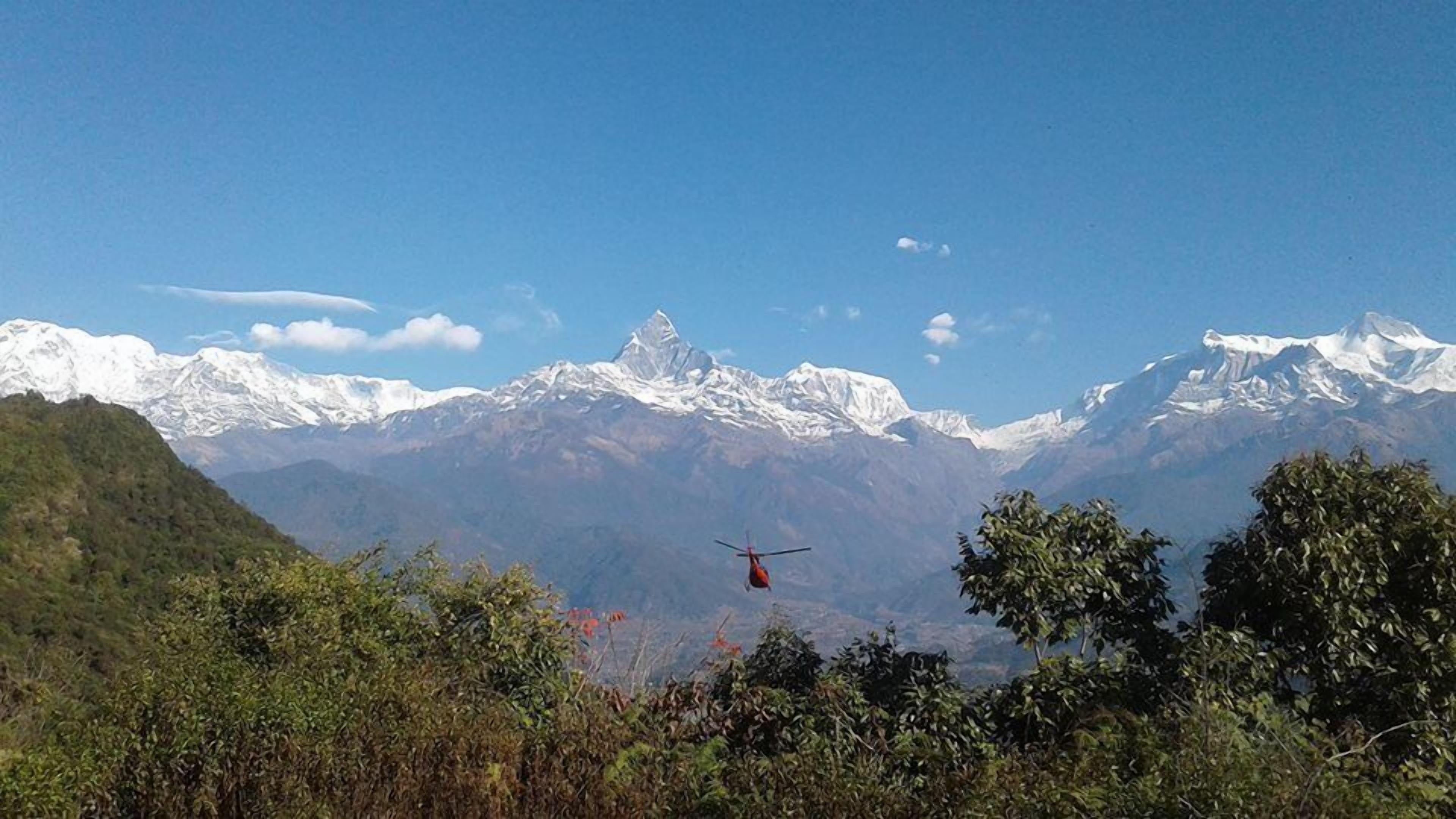 A helicopter flying over a mountainous landscape with snow-capped peaks and dense forest in the foreground.
