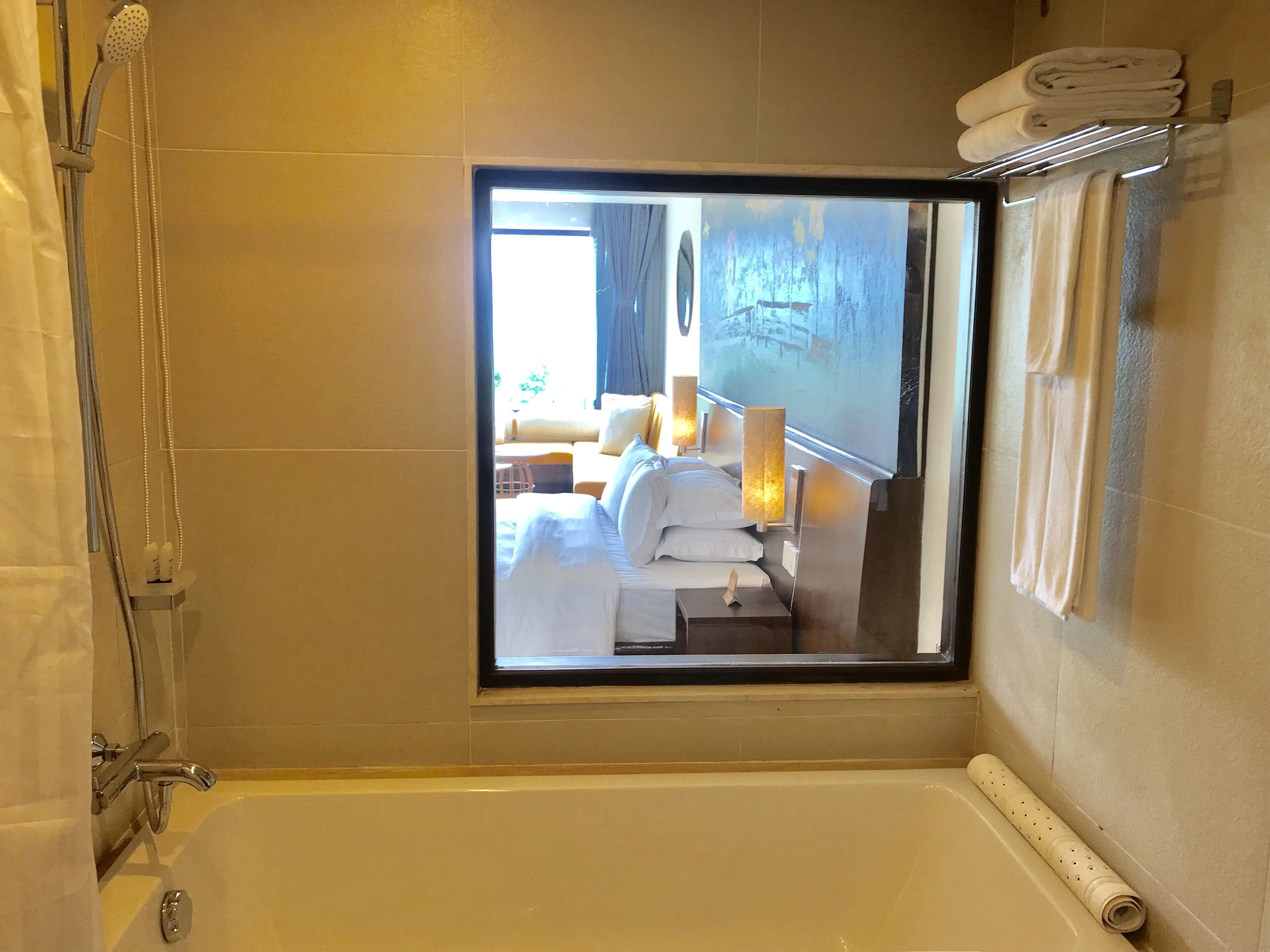 Une salle de bain d’hôtel équipée d’une baignoire, d’une douche et d’une fenêtre donnant sur une chambre avec un lit, une table de chevet et une lampe.